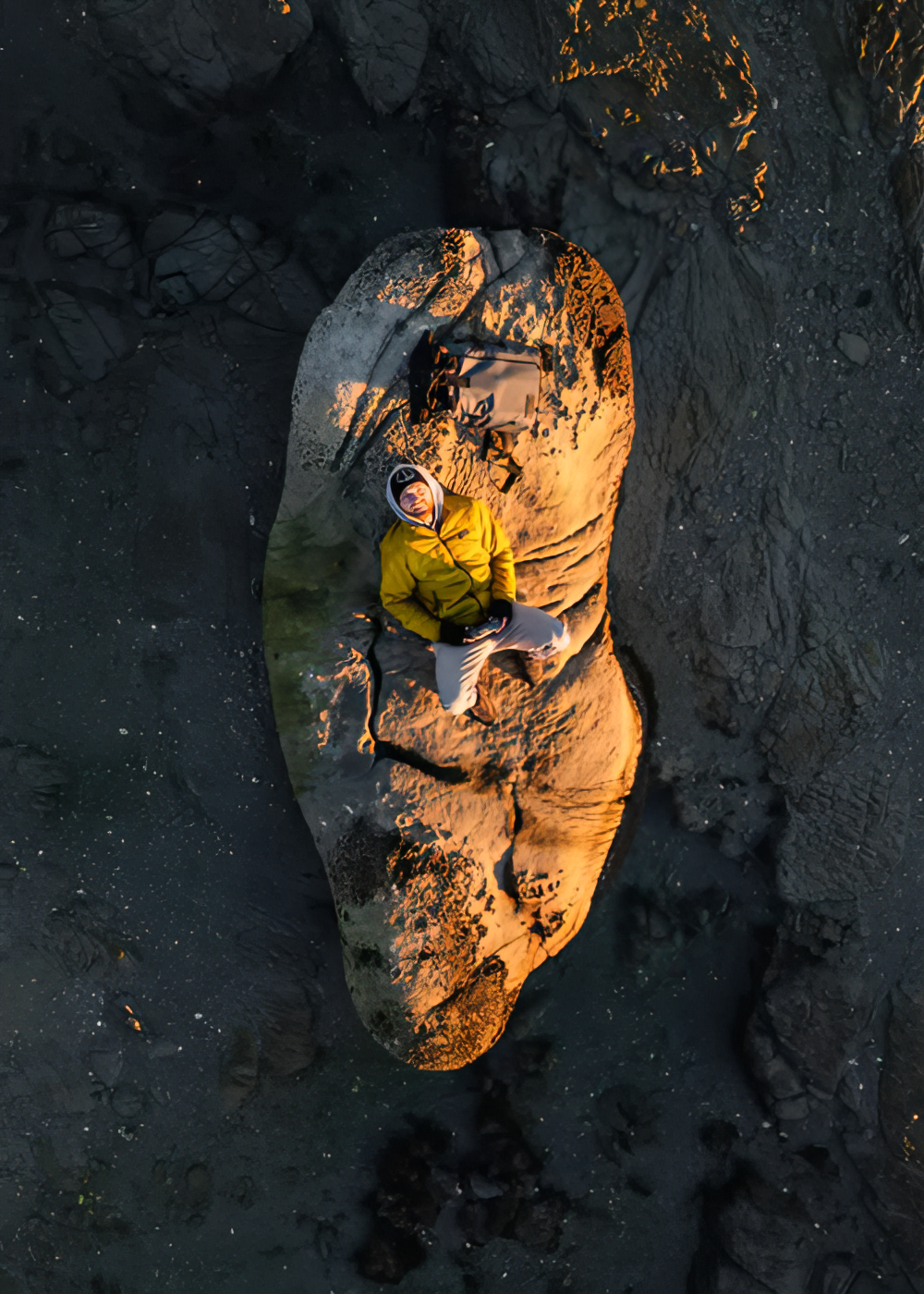 Person in gelber Jacke sitzt auf großem Felsen vor dunklem, strukturiertem Hintergrund